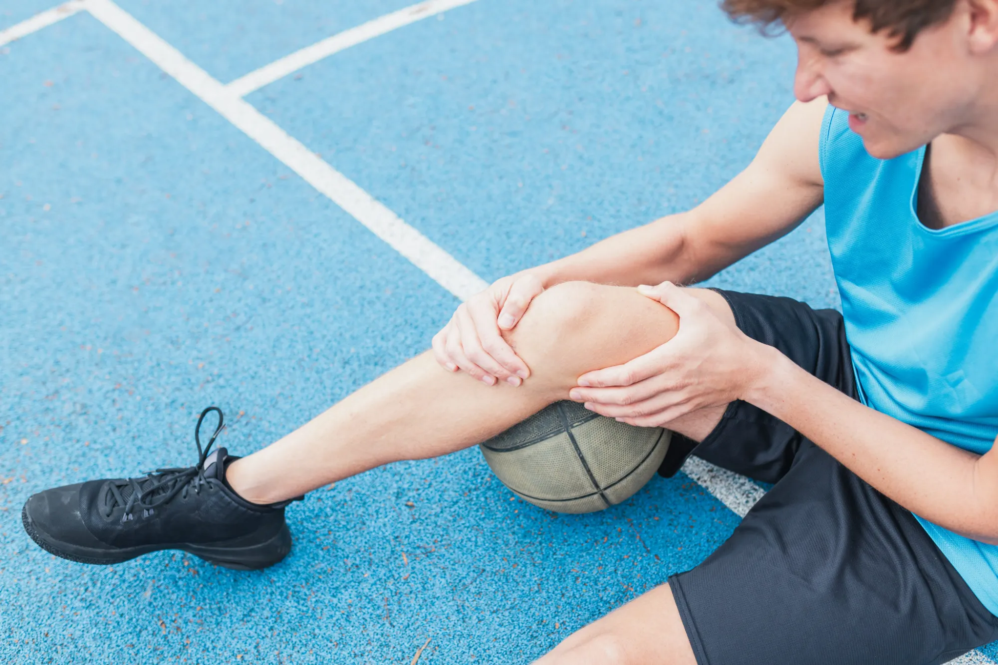 Young basketball player sitting on the court holding his injured knee